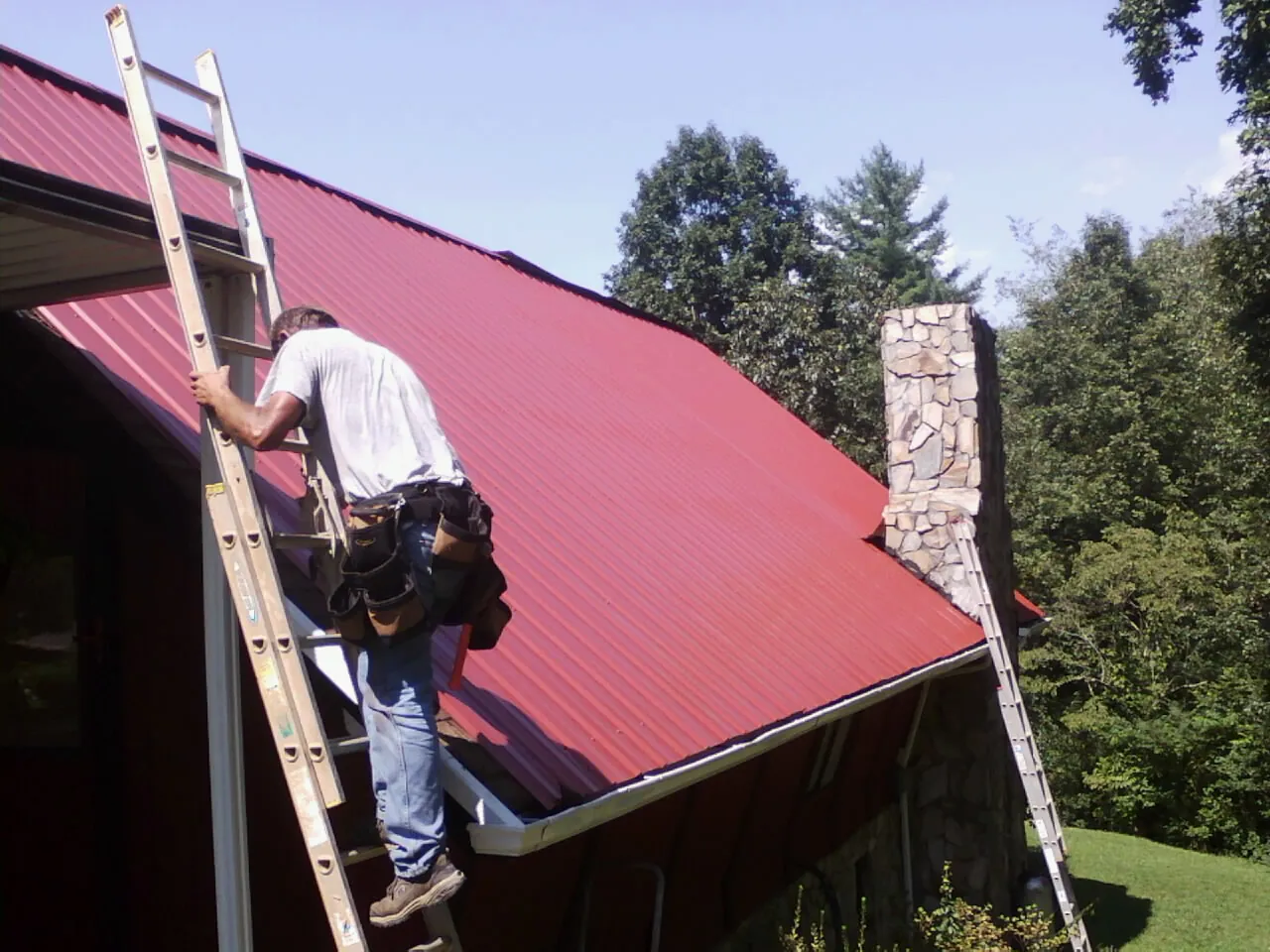 Roofer on a ladder beside a red metal roof and stone chimney — Soffit & Fascia Repair by Roofing Brothers in Keasbey